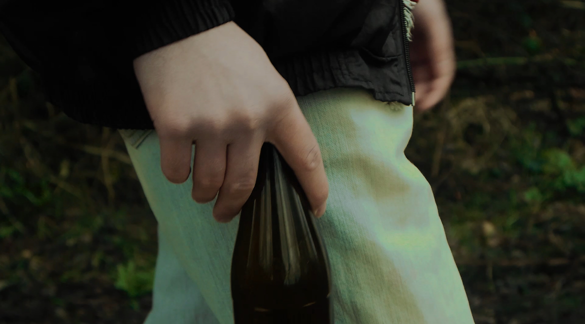 A hand holding a green glass bottle against pale denim in woodland
